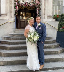 Bride and groom stand outside the ceremony with their bouquet of white peonies and the bride wears a headdress made of spray roses