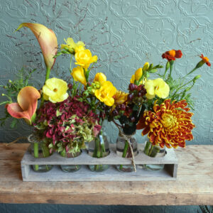 Photo showing Autumnal bottles, 5 small milk bottles in a wooden tray filled with autumnal seasonal flowers and foliages. Available to order from Kensington Flowers